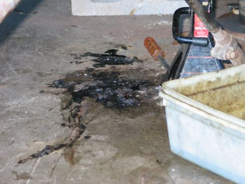 1995 Ford Bronco - Black tar-like sludge and spilled gear oil pooled on a concrete garage floor next to a drain pan, a screwdriver and a floor jack during a f…