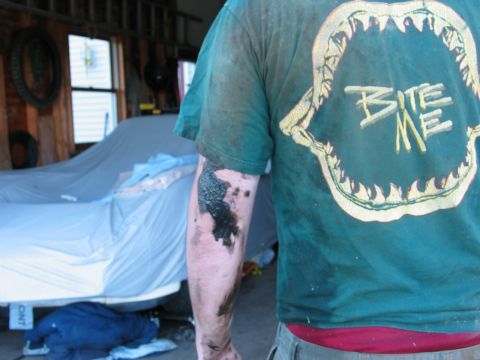 1995 Ford Bronco - Close-up of a person's forearm and back in a grease-stained green 'Bite Me' shark-jaws T-shirt inside a wood-walled garage, arm smeared wit…