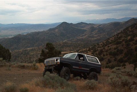 1986 Ford Bronco - A dark blue 1986 Ford Bronco with a white top is parked on a hillside overlooking a vast, mountainous landscape.