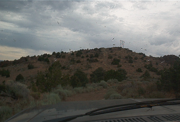 Dashboard and windshield view of a dry, hilly landscape with scattered bushes and a power line tower on the horizon