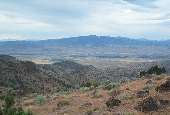 Wide view of a valley and mountains under a cloudy sky. Foreground has dry grass and rocks