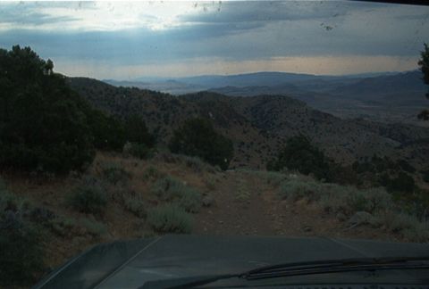 1986 Ford Bronco - View from inside a vehicle as it travels a dirt road through a vast, hilly landscape under a cloudy sky.
