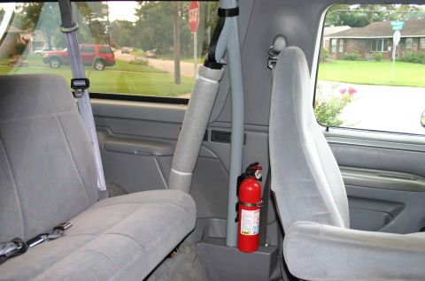 1994 Ford Bronco - Interior rear view of a Ford Bronco showing gray cloth seats, the gray B-pillar with mounted seatbelt, and a red ABC fire extinguisher secu…