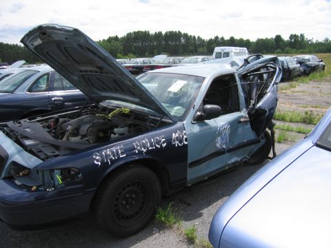 1991 Ford LTD Crown Victoria - A damaged state police car sits in a junkyard with its hood open and driver's side door mangled.