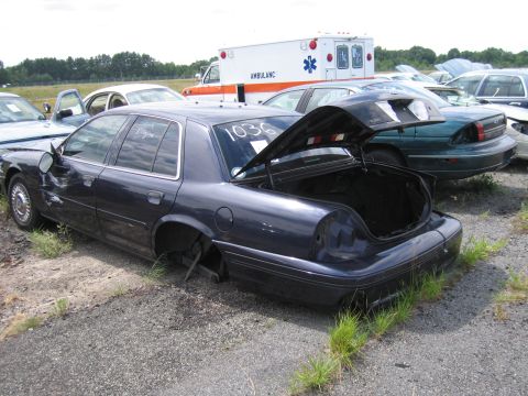 1991 Ford LTD Crown Victoria - A dark blue sedan sits in a junkyard with its trunk open and missing wheels, an ambulance visible in the background.