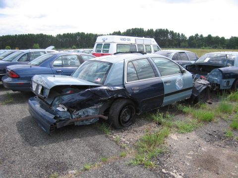 1991 Ford LTD Crown Victoria - A heavily damaged police car sits in a junkyard with other vehicles.