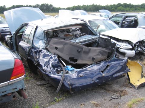 1991 Ford LTD Crown Victoria - A heavily damaged dark blue sedan sits in a junkyard with its rear end crushed and interior exposed.