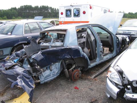 1991 Ford LTD Crown Victoria - A severely damaged blue sedan sits in a junkyard, its interior stripped bare, alongside other vehicles and an ambulance.
