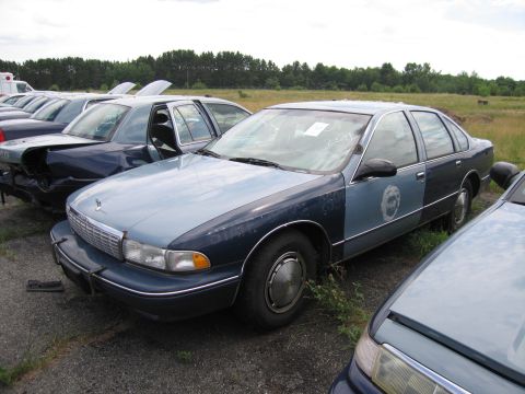 1991 Ford LTD Crown Victoria - A two-tone blue police car sits in a junkyard with other vehicles, showing a partial badge on its side.