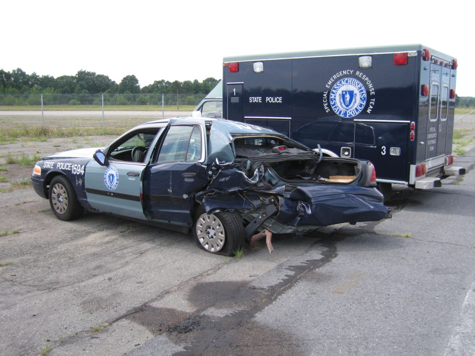 Rear view of wrecked 1991 Ford Crown Victoria LTD police car with flat tire and open trunk, next to an ambulance