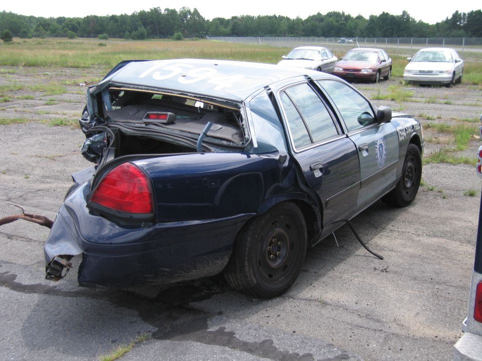 Damaged police car with trunk open, rear bumper crushed. Other cars in background