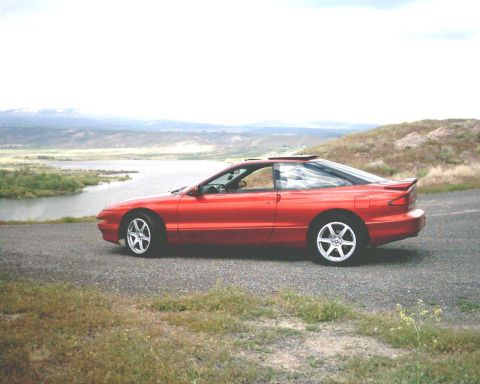 1996 Ford Probe - A red 1996 Ford Probe is seen from the side, parked on a gravel road with a scenic view of a lake and hills.