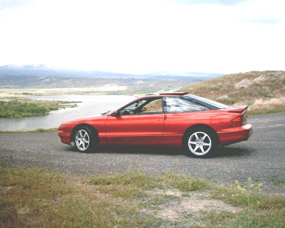 Red Ford Probe coupe, side view, sunroof open, at a river overlook