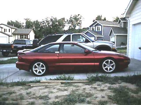 1996 Ford Probe - A red 1996 Ford Probe is parked in a driveway with other vehicles and houses in the background.
