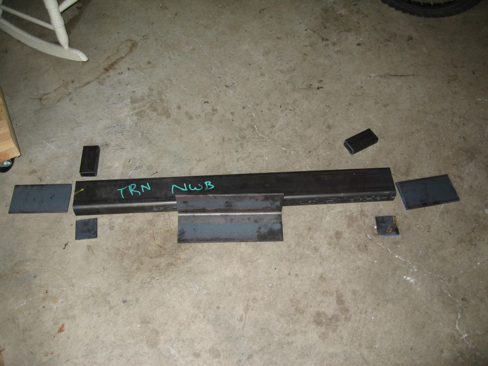 Steel tubing marked TRN NWB with angle iron and cut plates arranged on garage floor for fabrication