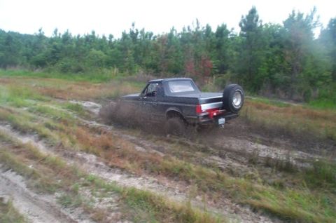 1988 Ford Bronco - A black 1988 Ford Bronco navigates a muddy off-road trail, its rear end partially submerged in the mire.