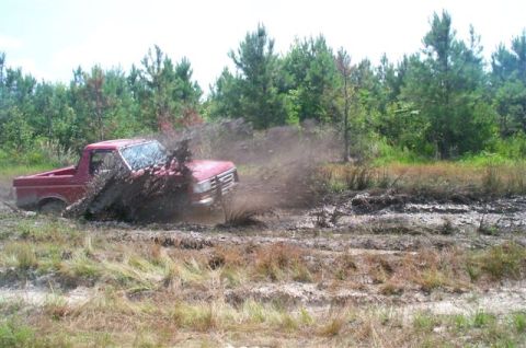 1988 Ford Bronco - A red pickup truck navigates a muddy terrain, sending a large spray of mud into the air.