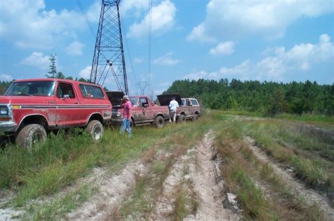 1988 Ford Bronco - Several vehicles are stopped on a dirt path next to a large power line tower, with people attending to their engines.