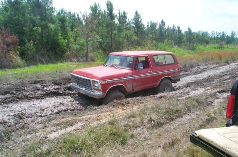 1988 Ford Bronco - This red SUV is stuck deep in the mud on a dirt track surrounded by trees.