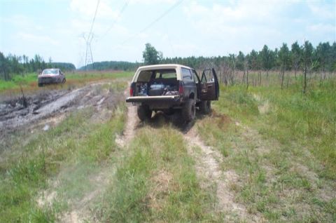 1988 Ford Bronco - Two vehicles are visible on a dirt track, one appears stuck in mud while the other is parked with its doors open.