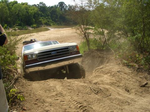 1990 Ford Bronco - A Ford F-Series truck navigates a challenging dirt path near a river.