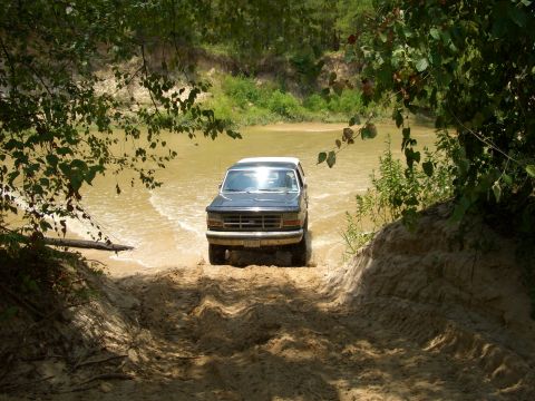 1990 Ford Bronco - A dark-colored Ford Bronco navigates through shallow, muddy water on a dirt path.