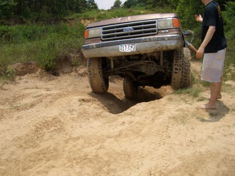 1990 Ford Bronco - A man stands by a muddy Ford F-Series truck as it navigates a sandy, off-road terrain.