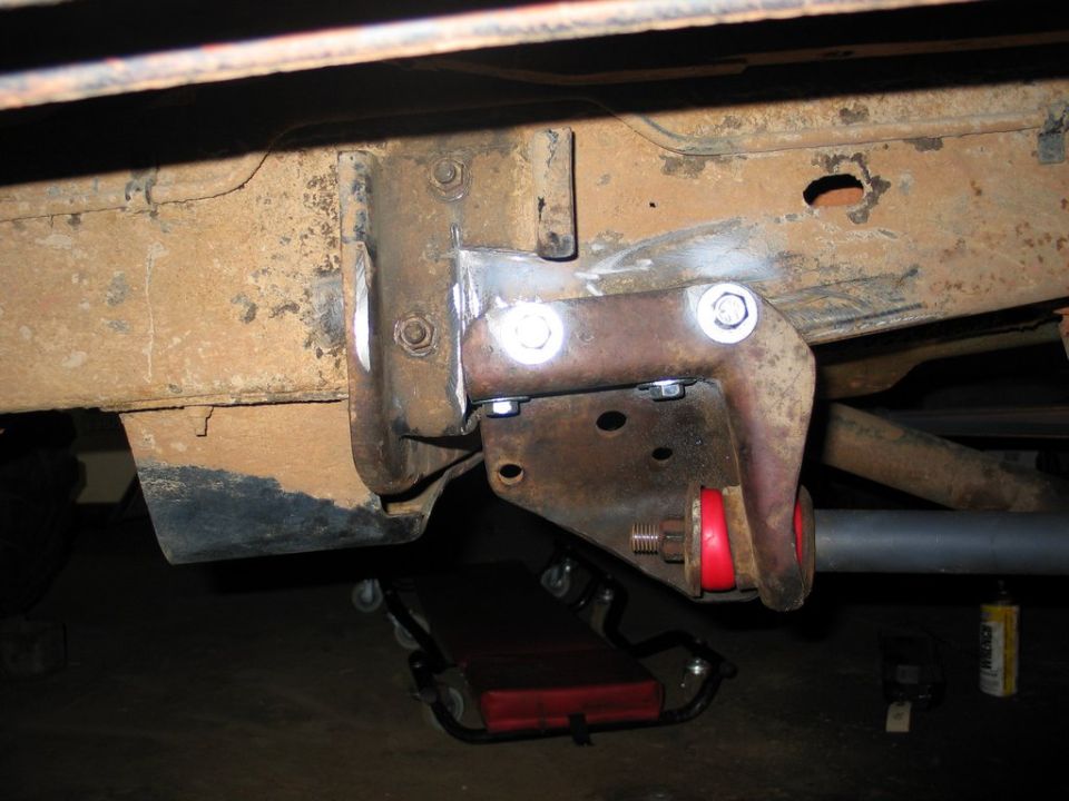 Underbody frame bracket with red bushing and silver bolts on a rusty truck chassis
