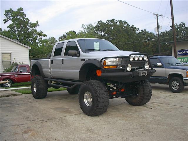 Lifted silver four-door pickup truck with large tires and a brush guard. Another pickup truck is parked behind it