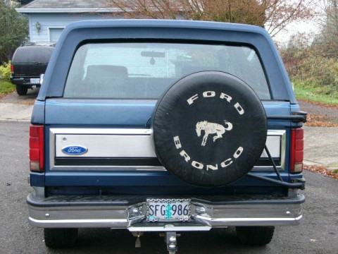 1986 Ford Bronco - Rear view of a blue 1986 Ford Bronco, a spare tire cover and a "SFG 986" license plate. Another vehicle is visible in the background.