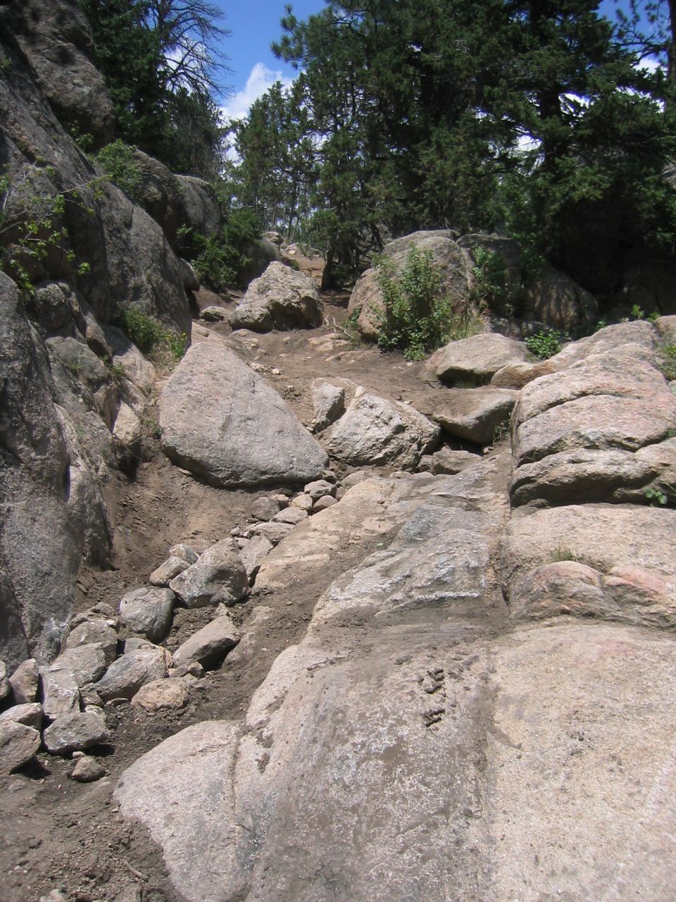 Rocky trail with large boulders and sparse trees under a blue sky. Text 'SUPERMOTORS www.supermotors.net' is visible on a rock