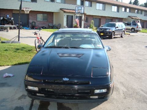 1989 Ford Probe - A black 1989 Ford Probe is parked in front of townhouses, with other vehicles and a bicycle nearby.