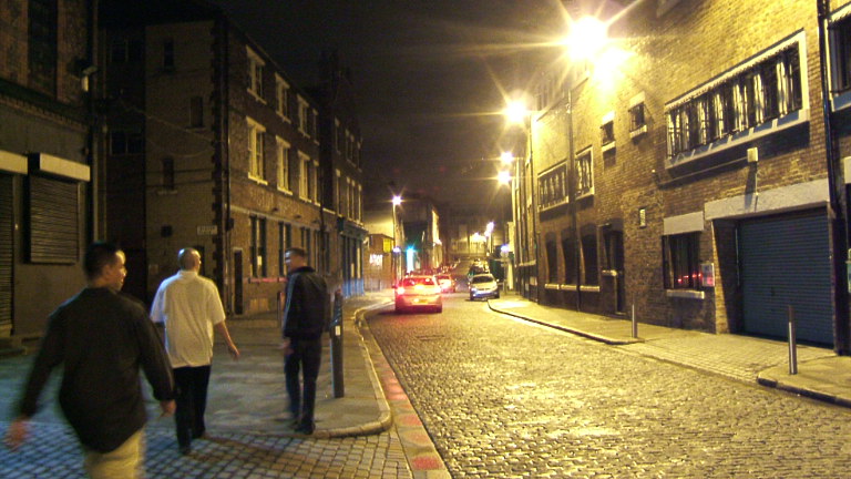 Three men walking a cobbled city street at night between brick buildings