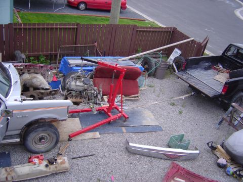 1986 Ford F-150 - A silver 1986 Ford F-150 is partially disassembled in a yard, with its engine on an engine hoist. A black pickup truck is parked nearby.