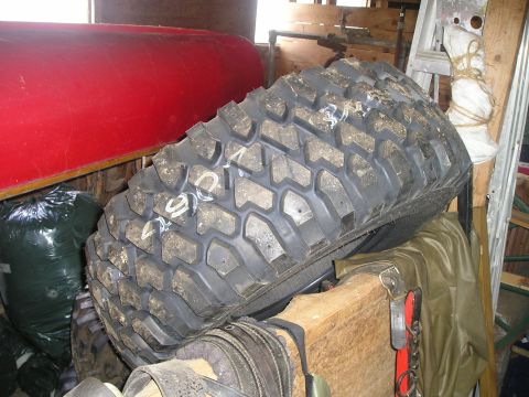 1984 Ford Bronco - A large, muddy off-road tire is stored indoors with other items, including a red canoe and a green bag.