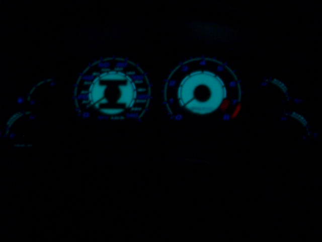 Glowing blue-green aftermarket gauge cluster at night showing speedometer and tachometer