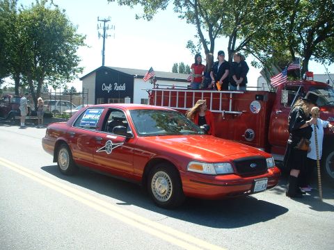 1999 Ford Crown Victoria - A red sedan participates in a parade alongside a fire truck, with people riding on the truck's bed.