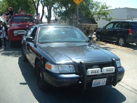 1999 Ford Crown Victoria - A Ford Crown Victoria police car is parked next to a red fire truck during what appears to be a parade.