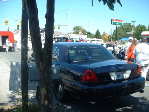 1999 Ford Crown Victoria - A dark blue sedan is parked on the side of the road, with people and other vehicles in the background.