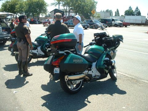 1999 Ford Crown Victoria - A green sheriff motorcycle is parked on the street, with two officers standing nearby. The motorcycle has saddlebags and a "SHERIFF" decal…