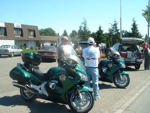 1999 Ford Crown Victoria - Two green police motorcycles are parked side-by-side with officers and civilians gathered around.