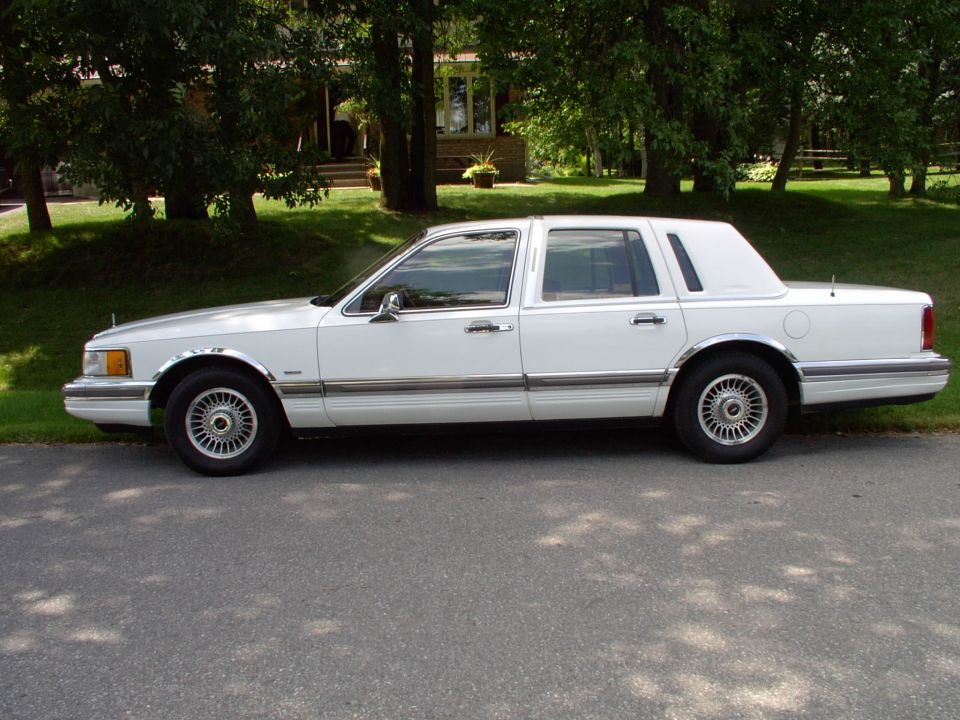 White Lincoln Town Car sedan in left side profile, parked on shaded driveway with trees and house behind