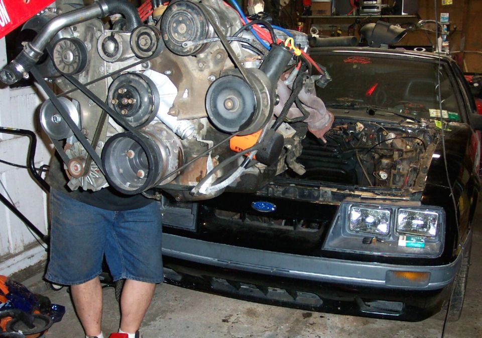 Man holding a V6 engine on a hoist over the open bay of a black Fox-body coupe in a garage