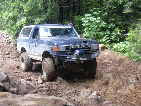 1991 Ford Bronco - A blue Ford Bronco navigates a challenging off-road path with large tires and auxiliary lights.