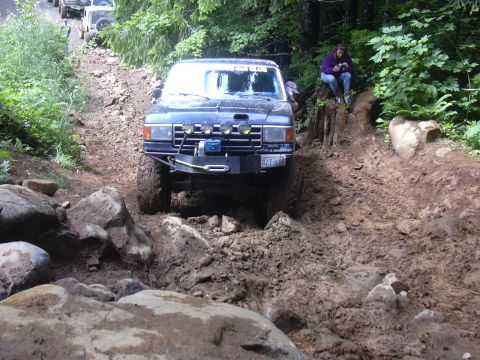 1991 Ford Bronco - Multiple off-road vehicles ascend a challenging muddy and rocky incline in a wooded area.