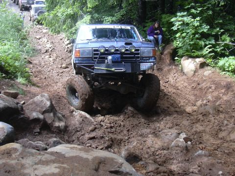 1991 Ford Bronco - An off-road SUV navigates a challenging muddy and rocky trail, with other vehicles visible further up the path.