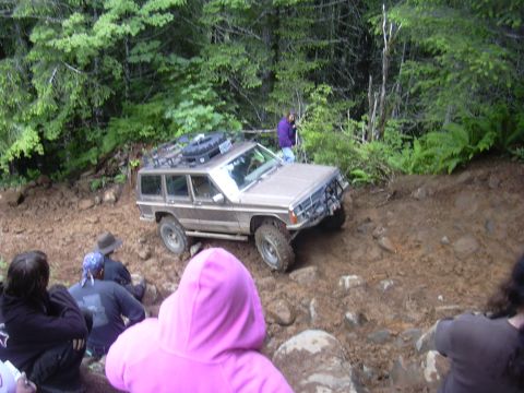 1991 Ford Bronco - A Jeep Cherokee XJ navigates a challenging muddy and rocky off-road trail, with onlookers observing the scene.