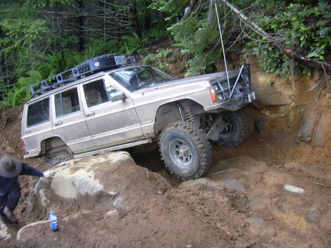 1991 Ford Bronco - A person guides the Jeep over a large rock on a steep, wooded incline.
