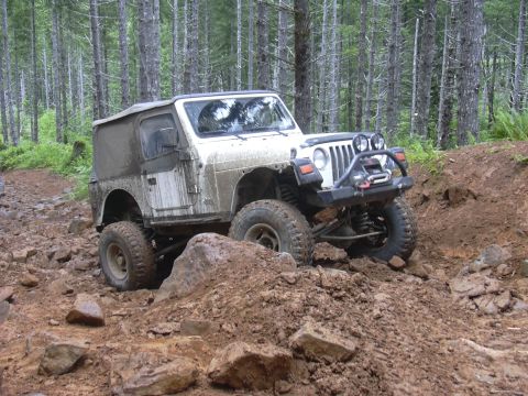 1991 Ford Bronco - A white Jeep Wrangler TJ navigates a challenging, muddy, and rocky off-road trail through a dense forest.