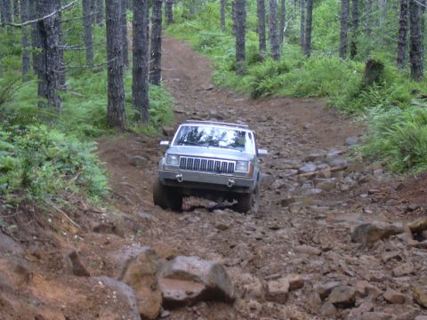 1991 Ford Bronco - A silver Jeep Cherokee XJ navigates a challenging, rocky path through a dense forest.